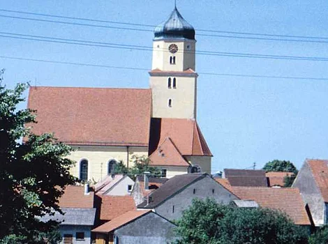 Kirchturm mit Uhr über Häusern mit roten Dächern und Bäumen vor blauem Himmel.