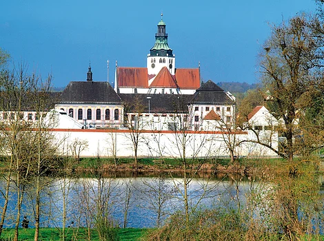 Klosteranlage mit Kirche und roten Dächern hinter Bäumen am Ufer eines Flusses bei klarem Himmel