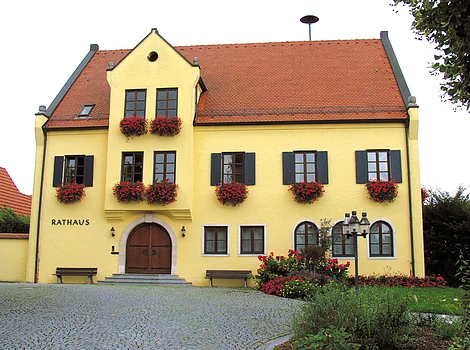 Gelbes Rathausgebäude mit roten Dachziegeln, Holzfenstern, Blumenkästen und gepflastertem Vorplatz.