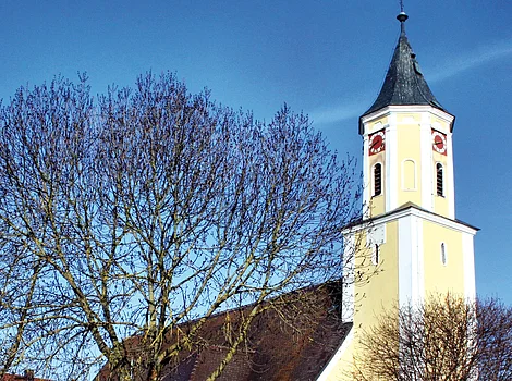 Kirche mit gelber Fassade und Turmuhr hinter kahlen Bäumen an einem sonnigen Tag