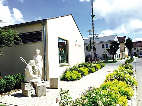 Skulptur mit zwei Figuren vor einem hellen Gebäude mit Fenster und Blumen entlang eines gepflasterten Wegs.