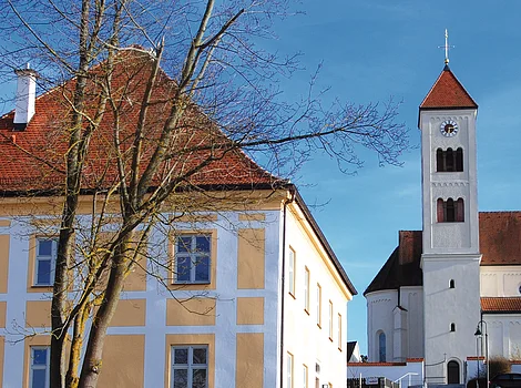 Gelbes Wohnhaus mit Baum davor und Treppe zu einer Kirche mit Turm im Hintergrund bei blauem Himmel.