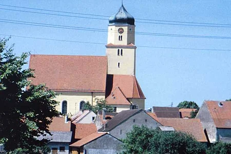 Kirchturm mit Uhr über Häusern mit roten Dächern und Bäumen vor blauem Himmel.