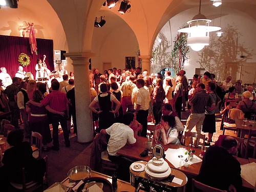 Menschen in einem Saal mit Rundbögen bei einer Veranstaltung mit Livemusik und Sitzplätzen an Tischen.
