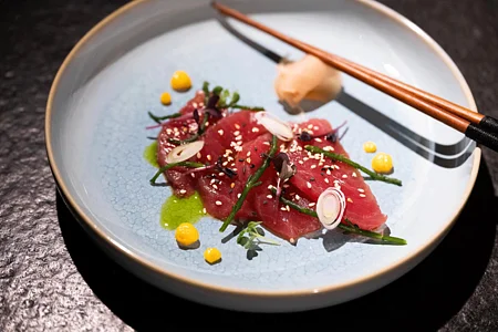 Dünne Scheiben rohen Fischs mit grünen Kräutern, Sesam, gelbem Mais und roten Zwiebeln auf einem schwarzen Tisch im blauen Teller mit Essstäbchen serviert.