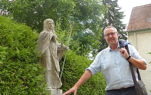 Der Monheimer Gästeführer Rainer Keßler Eine Person mit Jacke über der Schulter steht neben einer Statue in einem Garten, umgeben von Büschen und einem Gebäude im Hintergrund.