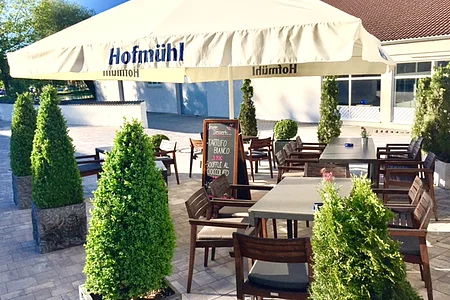 Außenbereich einer Pizzeria mit Tischen, Stühlen und einem Sonnenschirm umgeben von bepflanzten Kübeln auf gepflastertem Boden. Im Hintergrund ein Haus, Bäume und blauer Himmel zu sehen.