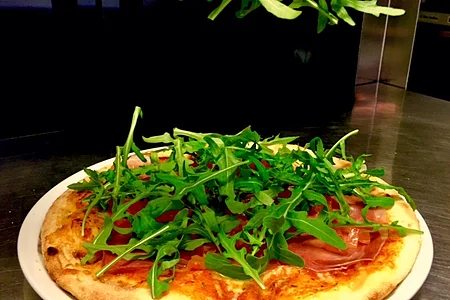 Hand legt Rucola auf eine belegte Pizza mit Schinken auf weißem Teller in Küche.