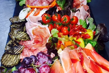 Antipasti-Platte auf schwarzer Unterlage mit Trauben, Tomaten, Melone, verschiedenen Wurst- und Käsesorten sowie gegrilltem Gemüse auf Salatblättern.