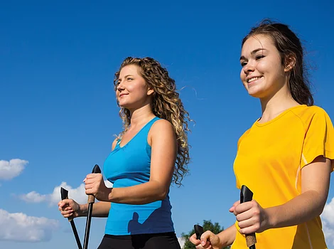 Zwei Frauen beim Nordic Walking auf einem Weg neben einer Wiese unter blauem Himmel mit Wolken.