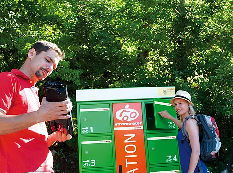 Stromtreter Mann hält Akku vor grün-orangefarbener E-Bike-Ladestation, Frau öffnet Fach, Fahrräder im Vordergrund.