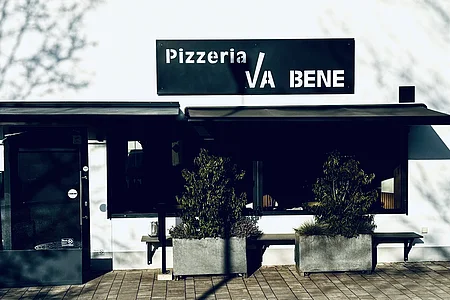 Außenansicht einer Pizzeria mit schwarzem Schild „Pizzeria VA BENE“, eine Eingansbereich aus Glas, verdunkeltem großen Fenster mit Rollläden und zwei großen Pflanzkübeln davor. Vor der Pizzeria ein gepflasterter Gehweg.