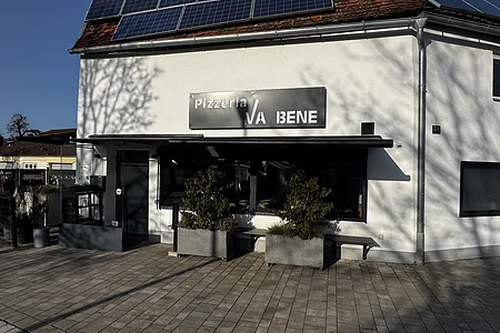 Außenansicht einer Pizzeria mit schwarzem Schild „Pizzeria VA BENE“, eine Eingangsbereich aus Glas, verdunkeltem großen Fenster mit Rollläden und zwei großen Pflanzkübeln davor. Vor der Pizzeria ein gepflasterter Gehweg. Auf dem Dach des  Gebäudes viele Solarpanele.