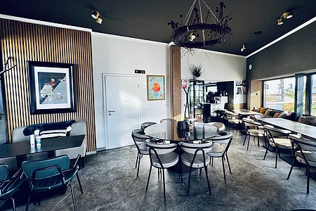 Moderner Innenraum einer Pizzeria mit Tischen, Stühlen, Wandbildern und einer Getränkekarte an der Wand. Größtenteils in grau-weiß eingerichtet mit Holzoptik-Fassaden an der Wand.