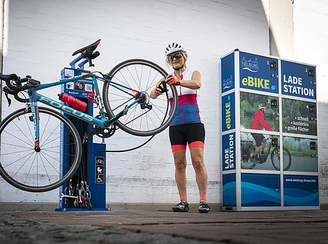 Frau repariert Fahrrad an blauer Reparaturstation neben eBike-Ladestation mit Werbebildern.