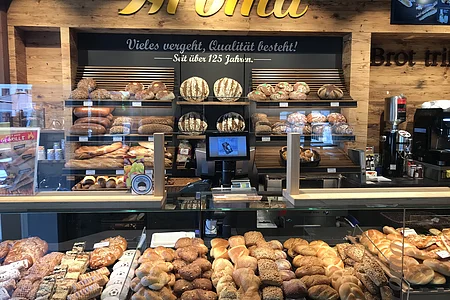 Bäckerei-Theke mit einer Vielzahl von Broten und Backwaren hinten auf Regalen und in Auslagen. An der Wand gelbe Aufschrift "Aroma".