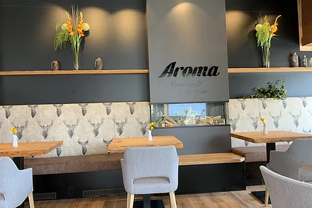 Innenraum eines modernen Cafés mit Holztischen, grauen Stühlen und einer Sitzbank mit Hirschmotiven. Oberhalb der Sitzbank stehen an einer schwarzen Wand Vasen mit Blumen.