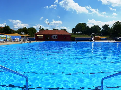 Außen-Schwimmbecken mit blauem Wasser, Umkleidehaus und Sonnensegeln bei klarem Himmel.