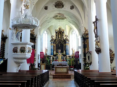 Innenraum einer Kirche mit weißen Säulen, Holzbänken und einem Altar am Ende des Mittelgangs