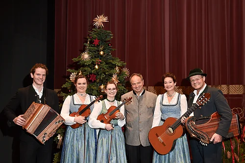 Sechs Personen in Trachten vor geschmücktem Weihnachtsbaum mit Musikinstrumenten posieren vor rotem Vorhang. Heilige Nacht mit Pfarrer R.M. Schießler & Familienmusik Rehm & Rupert Biegel