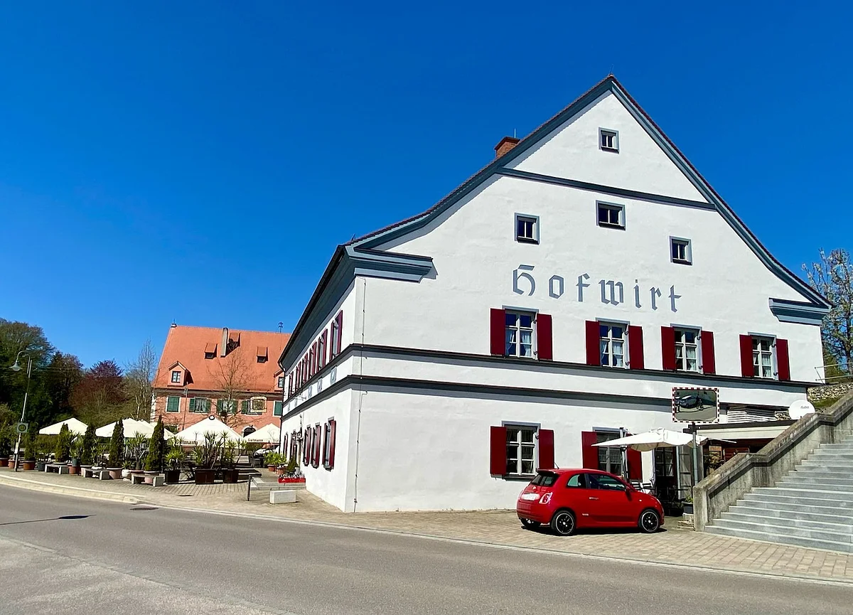Weißes Gasthaus mit rotem Auto davor und Außengastronomie bei klarem Himmel an Straße