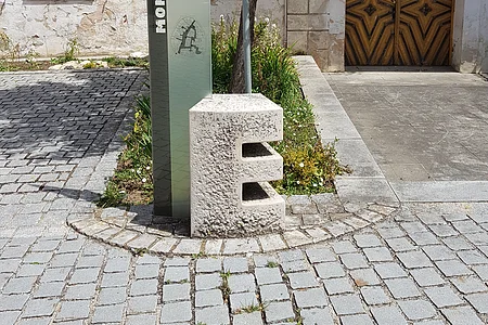 Steinskulptur des Buchstabens "E", dahinter ein Informationsschild und ein Baum mit einem dahinter liegendem Beet. Umgeben von einem gepflastertem Platz, mit einem historischen Gebäude im Hintergrund.