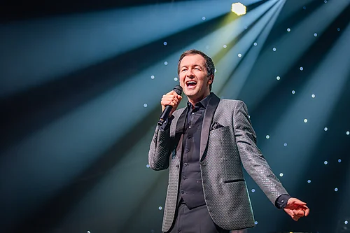 Mann im Anzug singt mit Mikrofon auf Bühne vor beleuchtetem Hintergrund mit Sternenlicht.
Udo Jürgens - Das Konzert mit Alex Parker & Special Guest am 20.02.2027 in der Monheimer Stadthalle.
