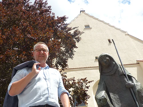 Der Monheimer Gästeführer Rainer Keßler Mann mit Jacke über der Schulter steht neben einer Statue, der Walburga-Statue in Monheim vor einem gelbgestrichenem Gebäude mit Giebeldach. Im Hintergrund ein großer Baum und sonniger Himmel.