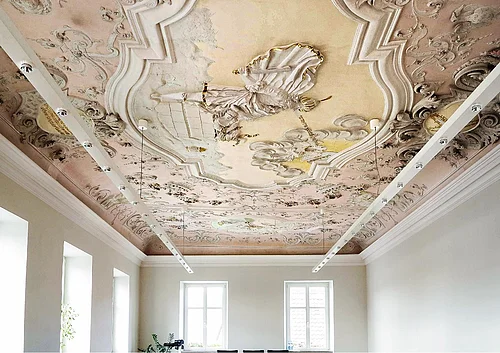Die Stuckdecke im großen Sitzungssaal - Abraham opfert Isaak, Isaaek segnet Jakob und Jakobs Traum Farbig gestaltete Decke mit barocker Stuck- und Freskoverzierung in einem hellen Raum mit weißen Wände, vielen Fenstern und moderner an der Decke hängender Beleuchtung.