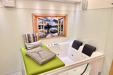 Whirlpool mit zwei Kopfstützen, grüner Sitzfläche, Handtuch und Fensterbild mit Bergsee-Motiv in weißem Raum