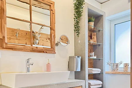 Badezimmer mit Waschbecken, Holzschrank, Spiegel, Handtüchern und Fenster mit Tageslicht.