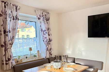 Essbereich mit Holztisch, vier Stühlen, Eckbank, gedecktem Tisch und Fenster mit gemusterten Vorhängen