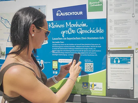 Startpunkt der Lauschtour "Kleines Monheim, große Geschichte" Frau mit Smartphone steht vor Infotafel zur Lauschtour in Monheim, Bayern, mit dem Titel "Kleines Monheim, große Geschichte" bei Sonnenschein. Frau hält ihr Smartphone an einen QR-Code.