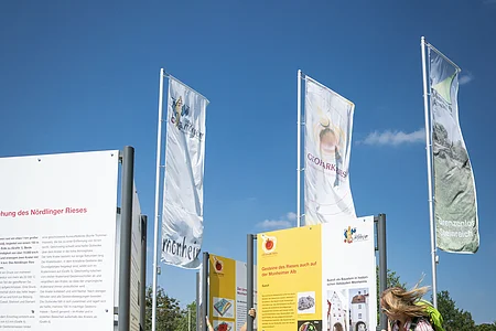 Eine erwachsene Personen und ein Kind stehen auf einer gepflasterten Fläche neben Informationstafeln im Freien, umgeben von Fahnen. Im Hintergrund grüne Wiese und blauer Himmel.