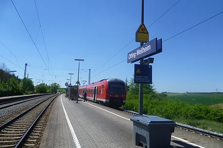Der Bahnhof Otting-Weilheim mit rotem Zug auf dem Gleis, blauer Himmel, grüne Landschaft im Hintergrund.