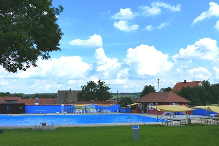 Freibad mit blauem Wasser, umgeben von Rasen, Bäumen und Gebäuden unter einem bewölkten Himmel. Vorderhalb vom Becken sind Sitzmöglichkeiten.
