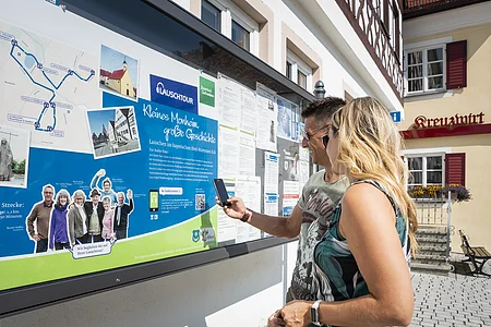 Zwei Personen vor einem weißen Gebäude, der Touristinformation Monheim betrachten eine Informationstafel mit einer Karte und Texten zur Lauschtour in Monheim und haben ein Smartphone in der Hand, mit dem sie einen Code abscannen.