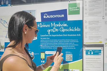 Frau mit Smartphone steht vor Infotafel zur Lauschtour in Monheim, Bayern, mit dem Titel "Kleines Monheim, große Geschichte" bei Sonnenschein. Frau hält ihr Smartphone an einen QR-Code.